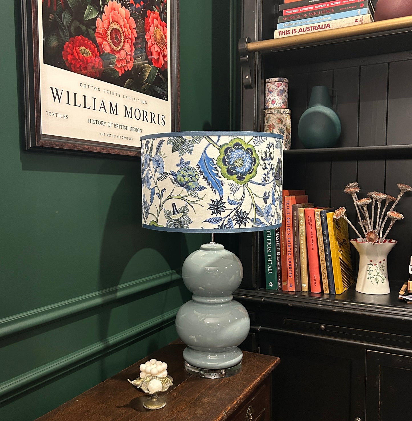 Decorative lamp with floral shade on a wooden table against a green wall with a framed picture.