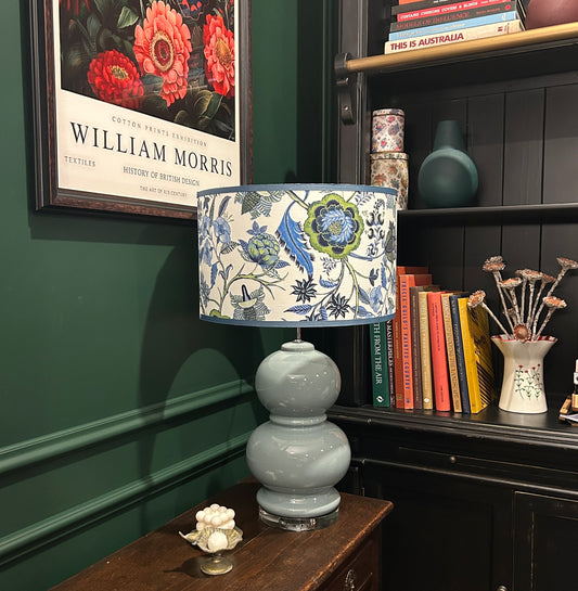 Decorative lamp with floral shade on a wooden table against a green wall with a framed picture.