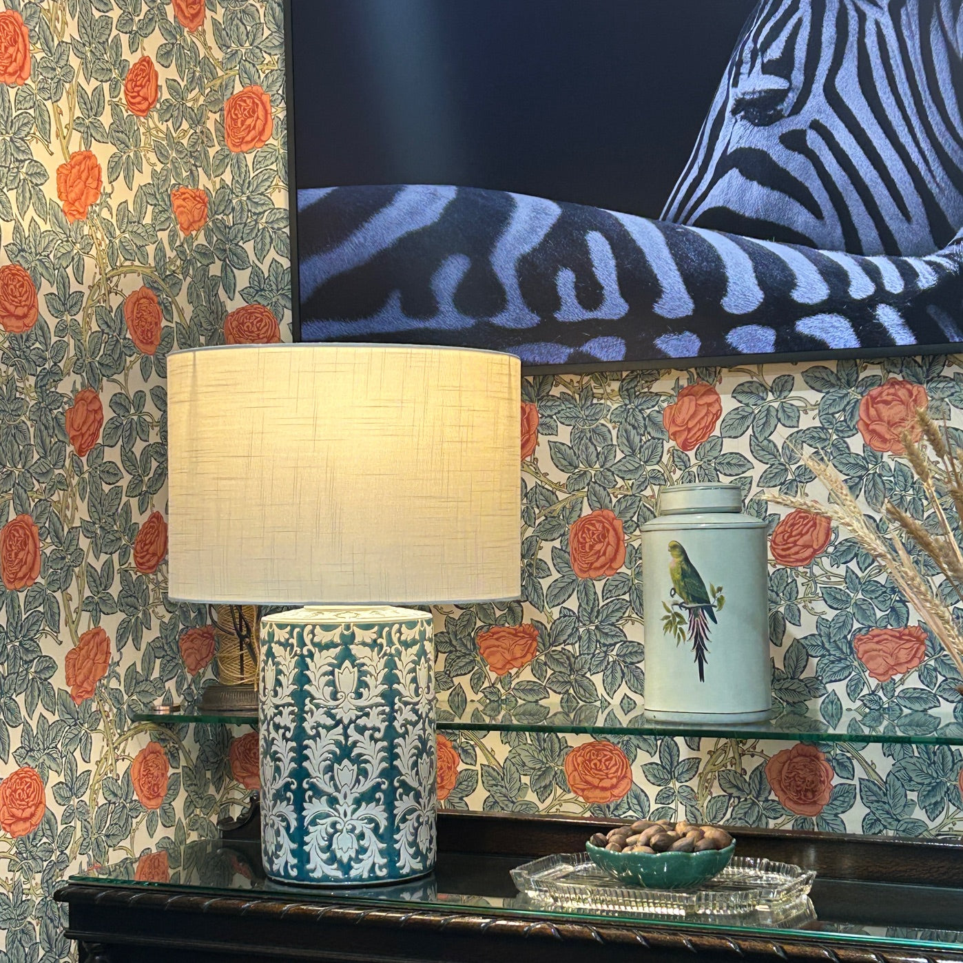 Decorative pattern teal green lamp with round drum lampshade featured against a floral wallpapered wall