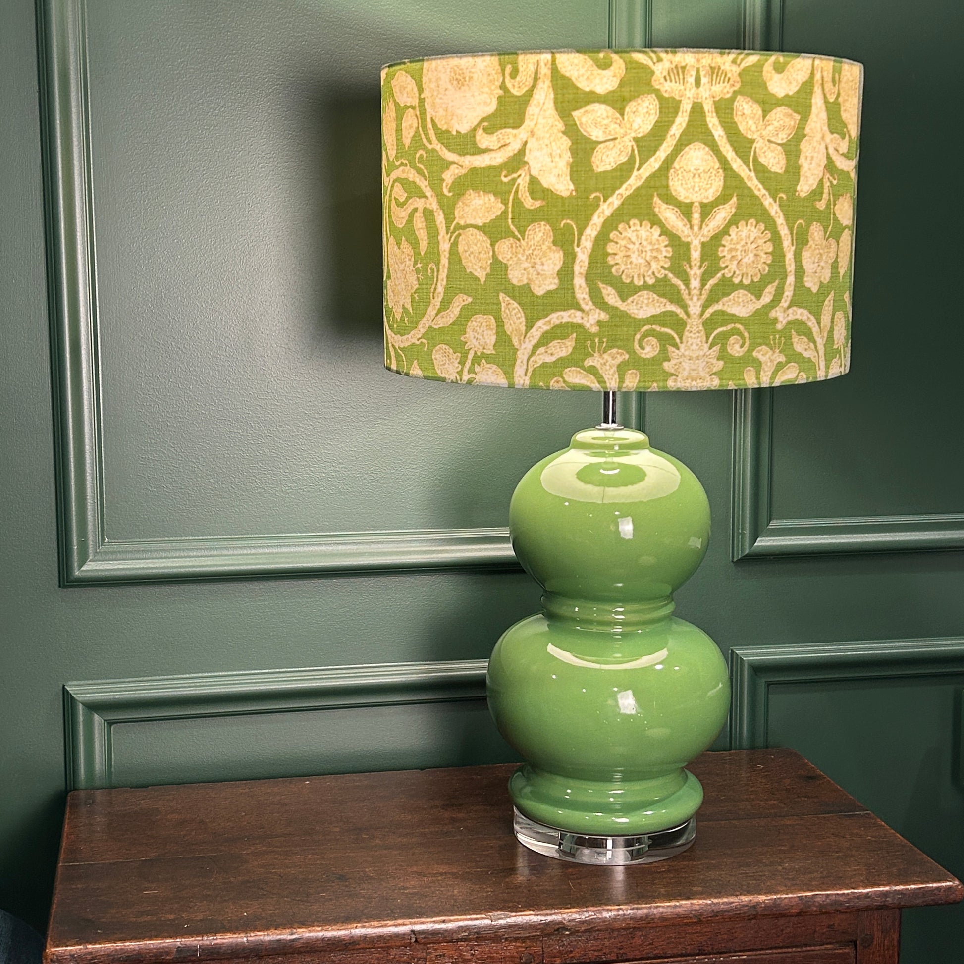 Green lamp with floral patterned shade on a wooden surface against a green wall.