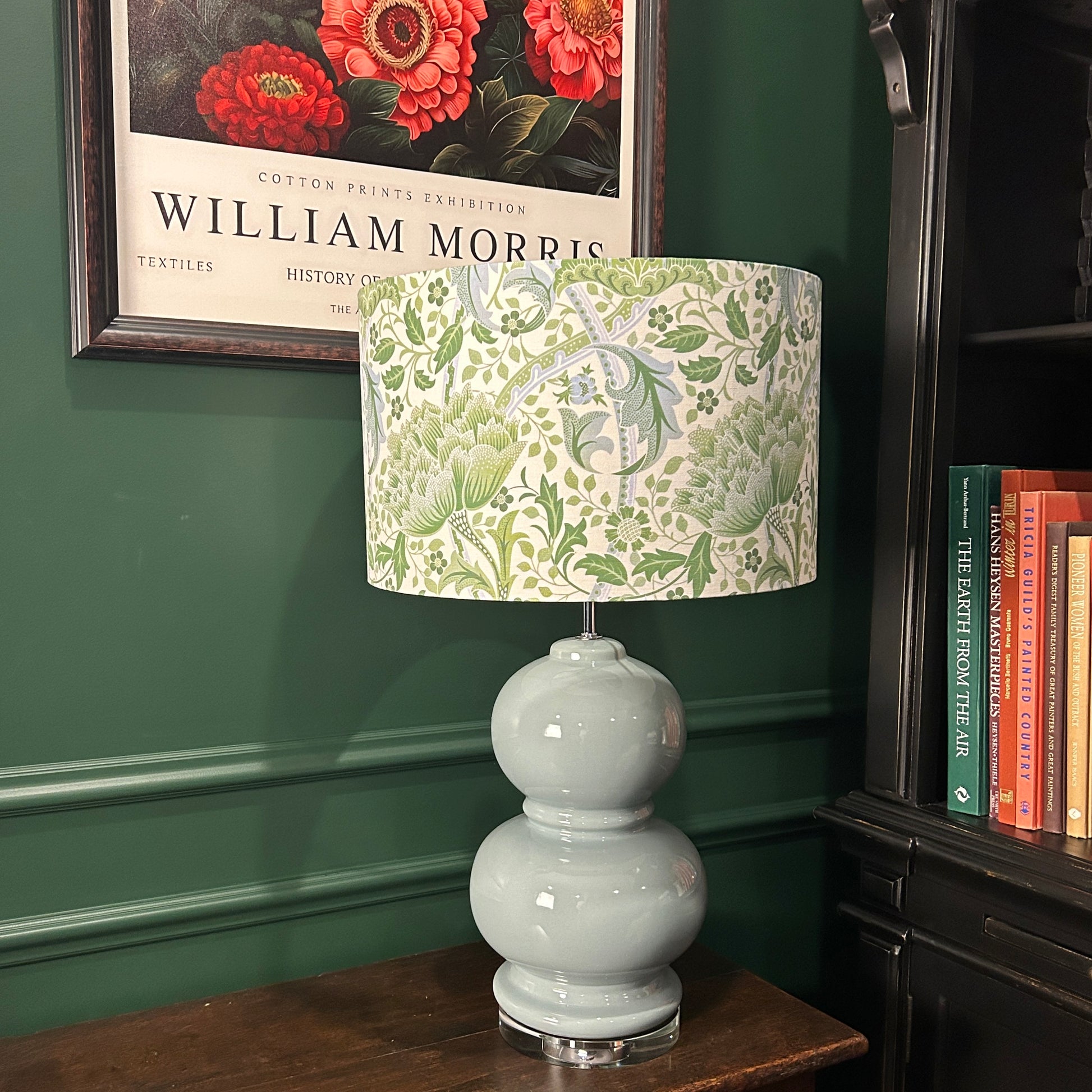 Decorative lamp with a floral patterned shade on a wooden surface against a green wall with a framed picture.